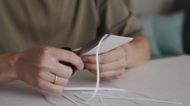 Close-up of hands cutting a curved strip from a sheet of white paper using scissors. Small paper pieces lie on the table during simple craft or office activity. High quality 4k footage
