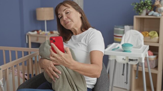 Woman holding smartphone and wrapped blanket beside a crib in a baby room inside a building; quiet concern.