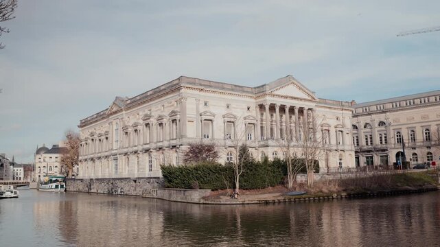 The grand neoclassical facade of the Ghent Palace of Justice rises above the Leie river bank, its elegant columns and triangular pediment reflected in the calm water under soft winter light.