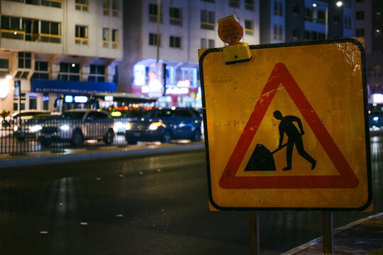 Roadwork warning sign with worker symbol on a busy street at night in Abu Dhabi, United Arab Emirates, with traffic lights and urban buildings blurred in the background.