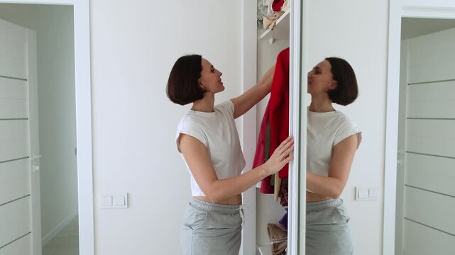 Woman Open the Closet and Choosing Red Outfit from Home Closet Rail &mdash; Stylish Brunette Holding Bright Garment While Decluttering Wardrobe in Modern White Interior. Wardrobe analysis