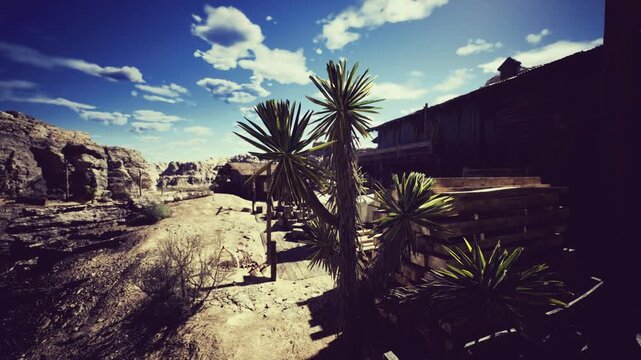 A rugged desert setting showcases vibrant green plants alongside wooden structures under a bright blue sky. The dry terrain is interspersed with rocky formations and scattered clouds.