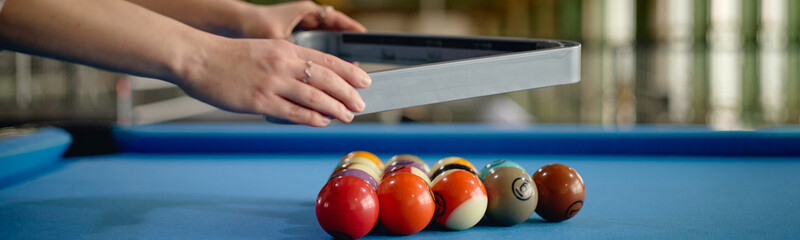 Website header shot of Caucasian young adult woman arranging billiard balls in triangle rack on blue pool table, hands visible positioning balls for game, indoor recreational setting, close up © Seventyfour