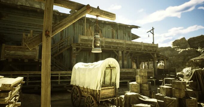 A rustic settlement showcases a wooden wagon draped with canvas, surrounded by crates and barrels. The bright sun highlights the rugged buildings in a western style setting.