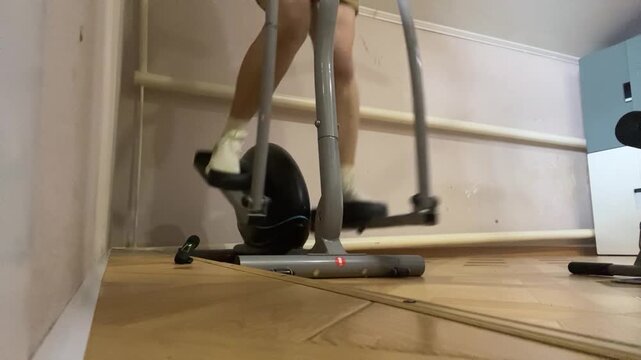 A person exercising on an elliptical machine indoors. The individual has light skin and is wearing white socks. The room features wooden flooring and gym equipment.