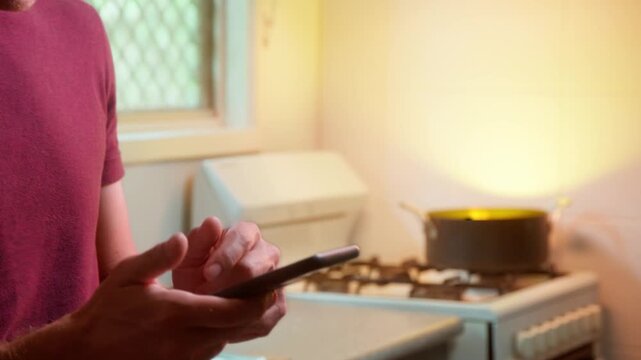Man in kitchen distracted by his phone fails to notice dinner in the saucepan behind him catching fire