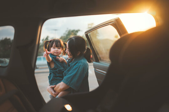 Happy Asian mother holding her young daughter by an open car door during sunset. Concept of family road trip, child safety, and emotional bonding. vacation journey