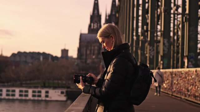 A beautifully composed sunset view from the Hohenzollern Bridge in Cologne, Germany. In the foreground, a woman photographer is seen from the side taking a picture.