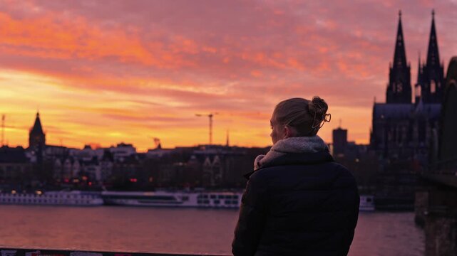 A woman seated on the Rhine River promenade, she looks across the river. She is the central subject, framed by the massive architecture of the Cologne Cathedral and the Hohenzollern Bridge.