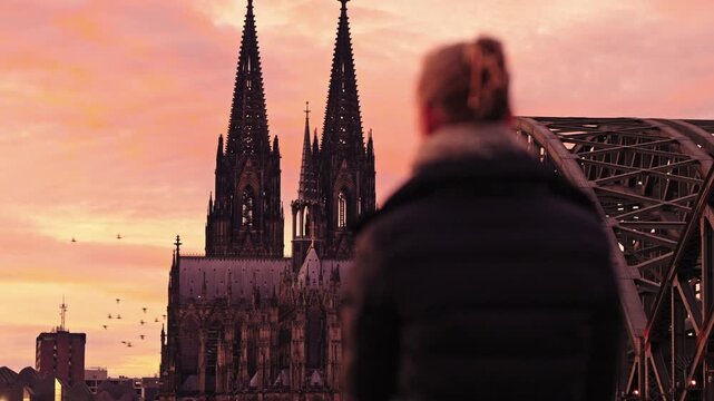 Woman admiring the iconic Cologne skyline from the Rheinboulevard in Deutz. The Hohenzollern Bridge and the Cologne Cathedral against a fiery sunset sky.