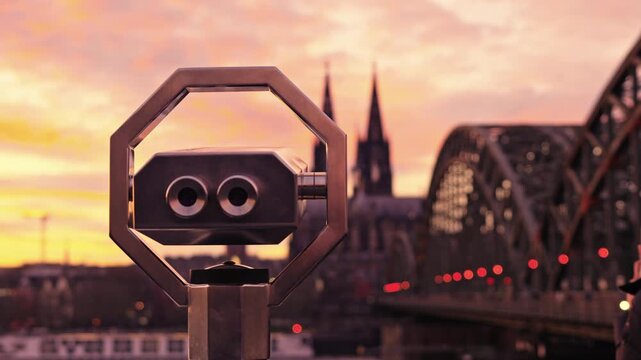 Vibrant sunset over the Rhine River in Cologne, featuring a woman filming the iconic skyline with the Hohenzollern Bridge and twin spires of the Cologne Cathedral.