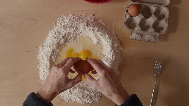 Overhead shot of a Romagna cook cracking multiple eggs into a flour well to prepare fresh cappelletti dough. Traditional Italian pasta-making with raw ingredients on a wooden table.