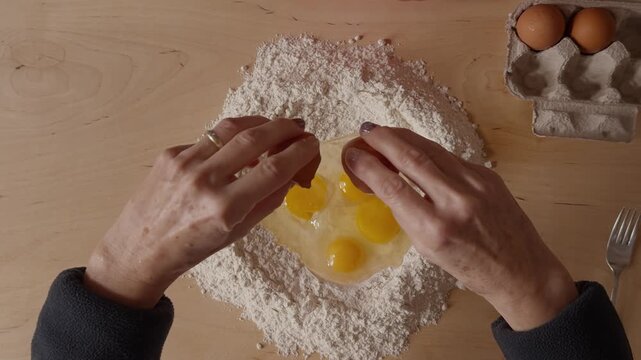 Top-down view of a Romagna cook cracking eggs into flour to prepare fresh cappelletti dough. Traditional Italian pasta-making with hands, ingredients, and rustic wooden table.