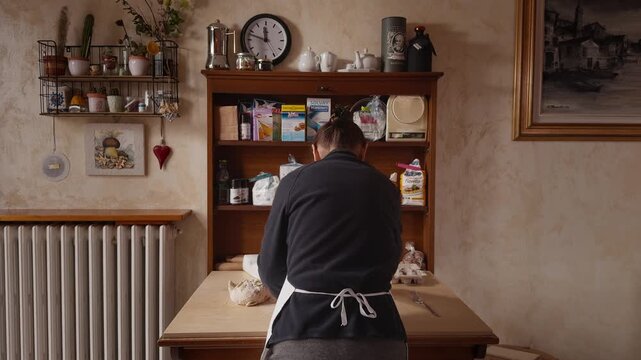 Rear view of a Romagna cook preparing fresh cappelletti dough in a traditional Italian home kitchen. Authentic pasta-making scene with wooden table, shelves, and domestic atmosphere.