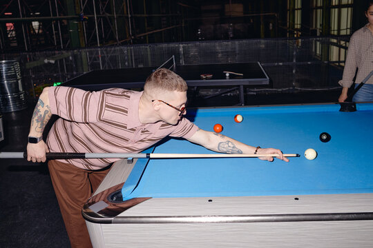 Caucasian young adult man with short hair and tattoos playing billiards, leaning over pool table while aiming cue stick at cue ball, another person partially visible in background