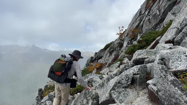 Ultralight backpacker navigates the steep, rocky slopes of Panorama Ginza Trail in the Hotaka Mountain Range. Fall foliage adds color to the rugged landscape under partly cloudy skies.