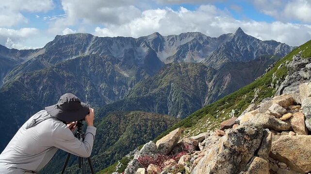 Middle-aged backpacker taking pictures of the rugged Hotaka Mountain Range on the Panorama Ginza trail in Nagano, capturing vivid fall colors under natural light.