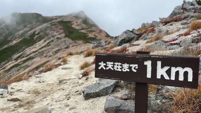 Woman hiker traverses rugged trail in Northern Japanese Alps, nearing Daitenso Hut in misty weather. Autumn hues and dynamic composition enhance the adventurous vibe.