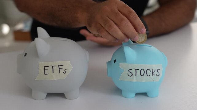 Close-up shot of hands inserting coins into labeled piggy banks. Gray ETF and blue STOCKS symbols under soft lighting. Financial planning, savings, and investment concepts highlighted.