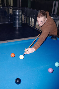 Vertical shot of young adult Caucasian man playing billiards, leaning over blue pool table while aiming cue stick at cue ball, focusing on shot with several colored balls visible on table