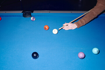 Caucasian young adult woman playing billiards, holding cue stick and preparing to strike cue ball on blue pool table, several colorful balls scattered across surface