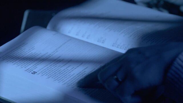 A detailed close-up shot of hands engaged in reading a Bible under cool, low lighting, creating an intimate and contemplative atmosphere. The image captures focus and calm.