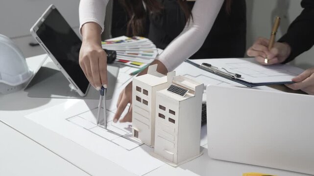 Architects reviewing building plans with scale model, laptop and tablet on desk, hands drawing and taking notes in bright modern office.