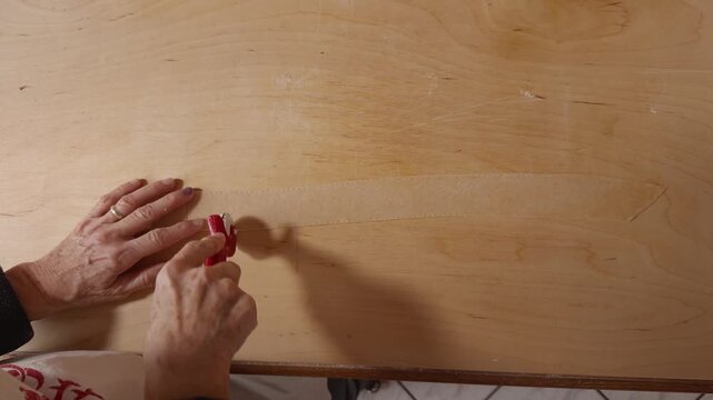 Top-down view of elderly hands using a red pasta cutter to slice dough into small squares on a wooden surface.