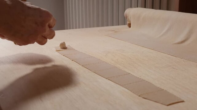 Hands adding ricotta filling to small pasta squares laid out on a wooden surface, preparing homemade cappelletti.