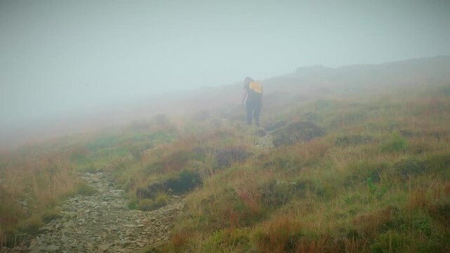Soft mist drifts across rolling highland meadow with distant peaks barely visible through haze, hiker navigates trail