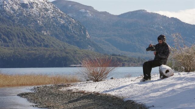 A young man in a winter jacket sits on a snow-covered log, pouring a traditional mate drink with heated water on thermos, beside a tranquil glacial lake in the mountains of Patagonia, Argentina