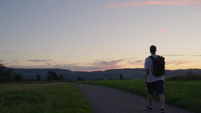 Young man with a backpack walking down on a footpath at sunset in summer, Switzerland