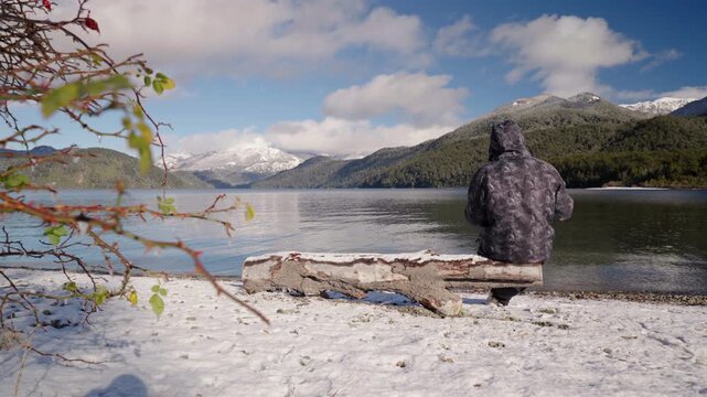 Man relaxes sitting on log by snowy lake sipping yerba mate in Patagonian winter mountains, Argentina, static rearview with space for text