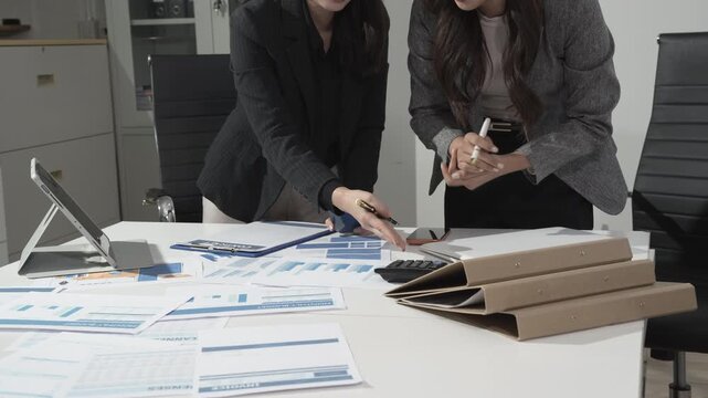 Two businesswomen in an office lean over a desk covered with financial reports, calculator and folders, collaborating on analysis and planning.
