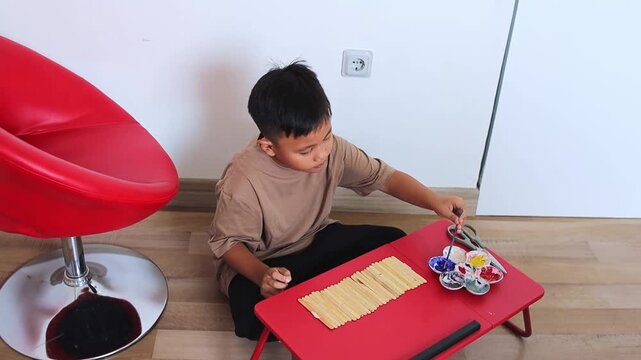 Creative kid enjoying DIY art project painting wooden sticks using colorful paints on folding table in room