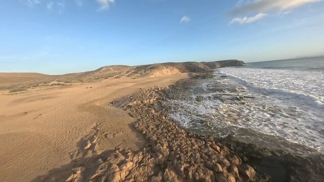 Aerial view of sunset over sandy beautiful beach and ocean. FPV drone flying over sand and water, birds eye view.