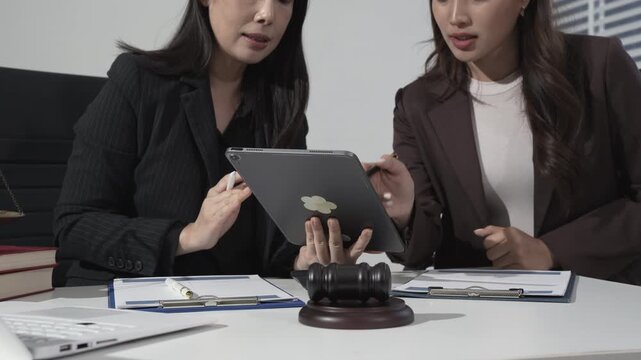 Two businesswomen in suits review a tablet at a desk with legal documents and a judge gavel, suggesting a law consultation or corporate compliance meeting in a modern office.
