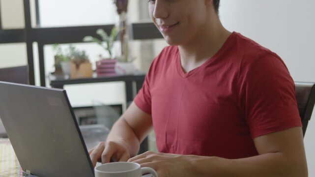 Young man types on laptop at a table with a coffee mug. He seems happy and focused on his tasks as he chuckles positively - Tilt up - Slow Motion