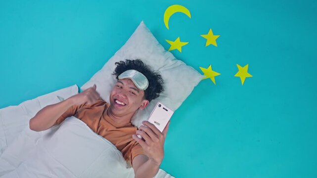 Top view of smiling man relaxing in bed holding mobile phone and gesturing thumb up on blue studio set