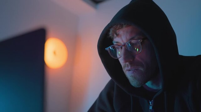 Cybersecurity hacker in hoodie using comp in hoodie using computer at night with screen glow on his face. Cybersecurity, hacking, cybercrime, data breach, malware, phishing, and online threat concept.