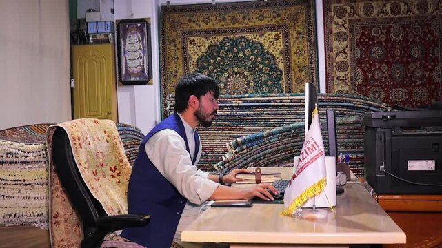 A man in Kabul city is selling rugs in large quantities in carpet shop behind computer screen. many carpets on background, Afghanistan economy and modern office work meets ancient culture concept