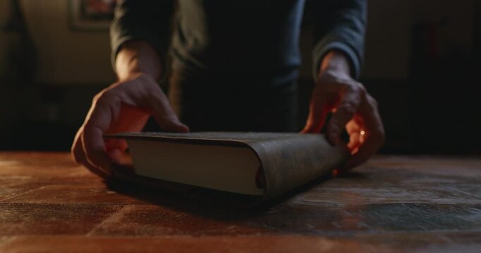 A person carefully opens a large book on a wooden table, focusing on the texture and details of the cover.