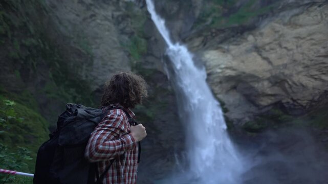 Backpacker hiking close to a powerful alpine waterfall in the Swiss Alps, pausing to observe flowing water and steep rock walls. Reichenbach waterfall near Meiringen, dramatic nature outdoor exploring