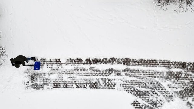 Drone top-down shot of a person clearing a cobblestone driveway with a blue shovel, revealing brick pattern against fresh white ground with wide copy space.