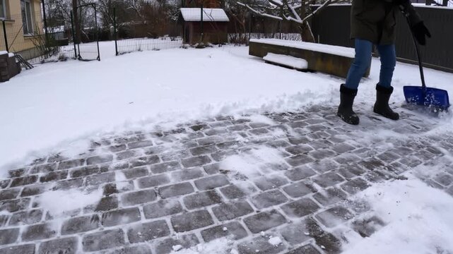 Wide low-angle shot of a person pushing a blue shovel across a cobblestone patio, sweeping powder aside and revealing wet pavers during backyard winter cleanup.