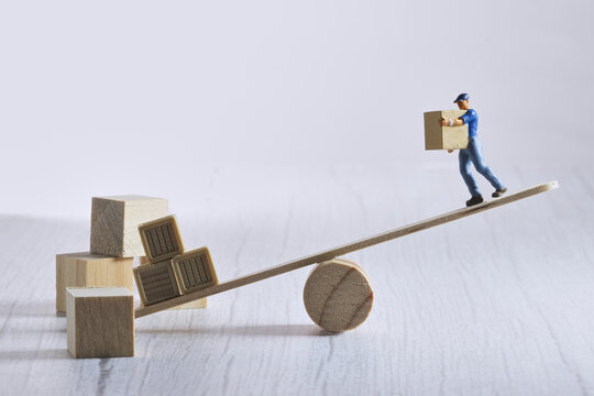 Miniature worker carrying a box on an unbalanced seesaw structure