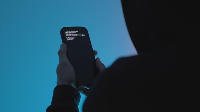 Sensitive data theft. Hooded person holding smartphone with green code on screen in blue light. Hacking, cybercrime, malware, data breach, and cybersecurity threat concept. Virus in smart phone,