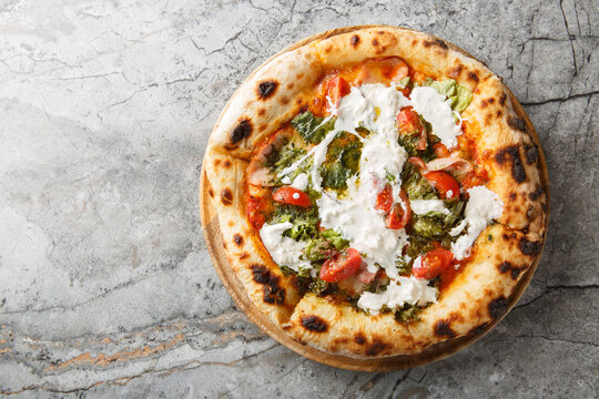 Delicious authentic pizza with stracciatella cheese, pesto, ham and cherry tomatoes close-up on a wooden board on the table. Horizontal top view from above