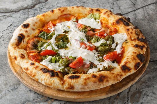 Delicious authentic pizza with stracciatella cheese, pesto, ham and cherry tomatoes close-up on a wooden board on the table. Horizontal