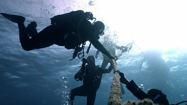 Scuba divers pause midwater while holding a descent line during a safety stop, silhouetted against bright surface light as streams of bubbles rise through the deep blue sea.